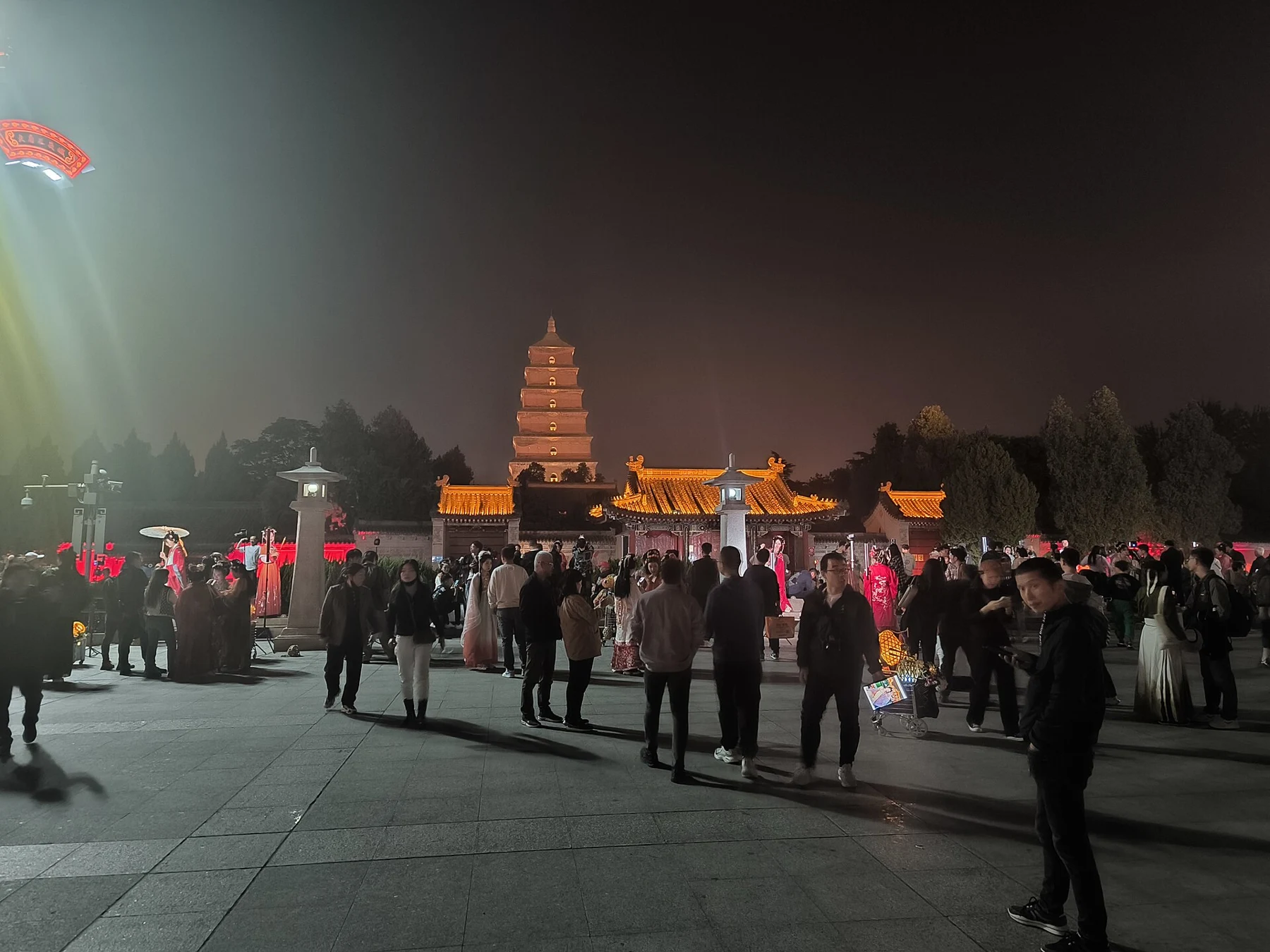 People crossing an open square, with a lit tower rising beyond a roofed entrance.