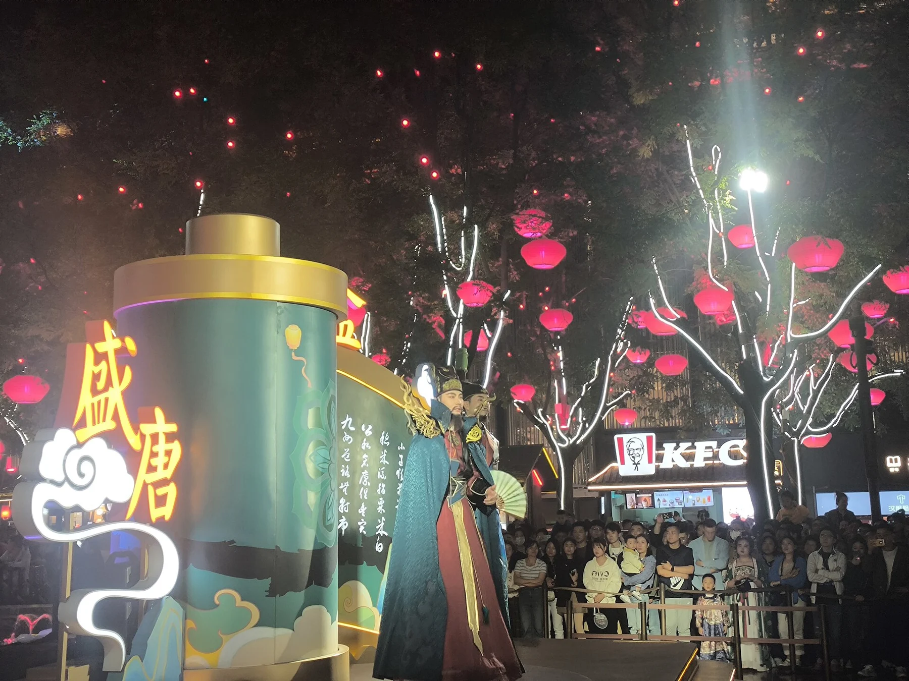 A costumed figure standing next to a green cylindrical prop in a lantern-lit street.