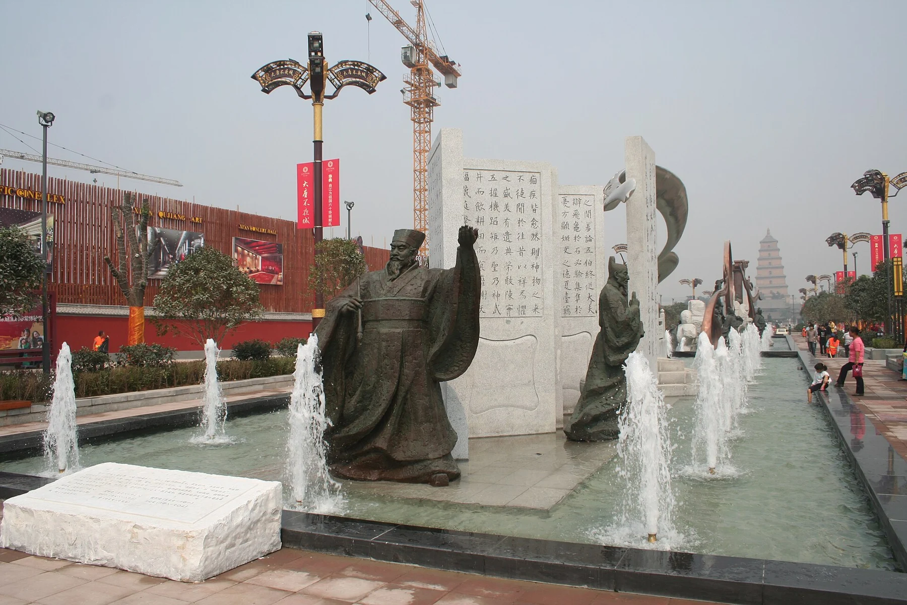 Robed sculptural figures standing beside a tall inscribed stone and small fountains.