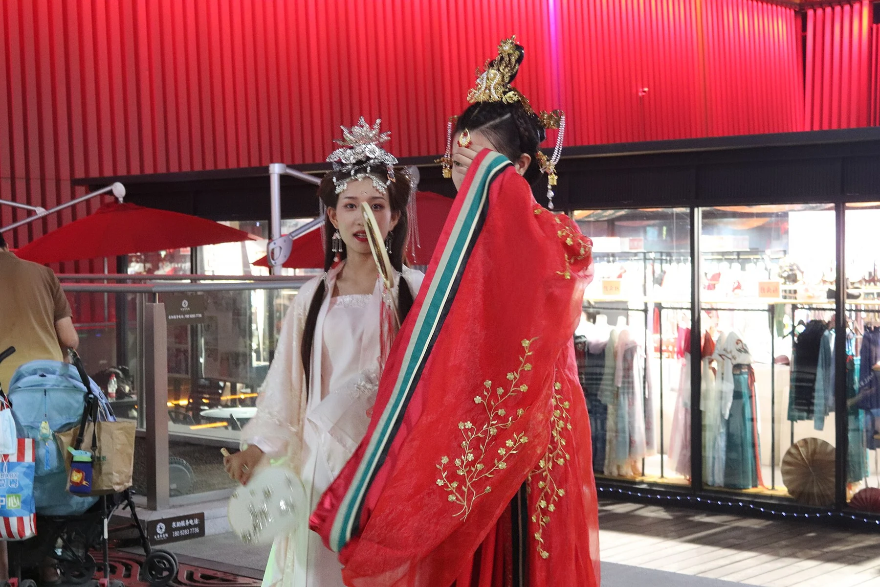 Two people in ornate red-and-white costume standing beside a contemporary shop facade.