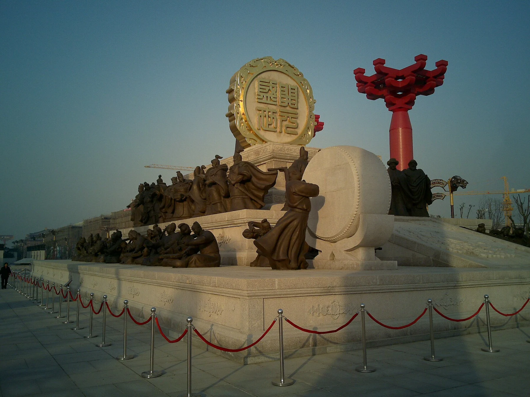 Seated sculptural musicians below a large round panel, viewed from the side of a pale platform.