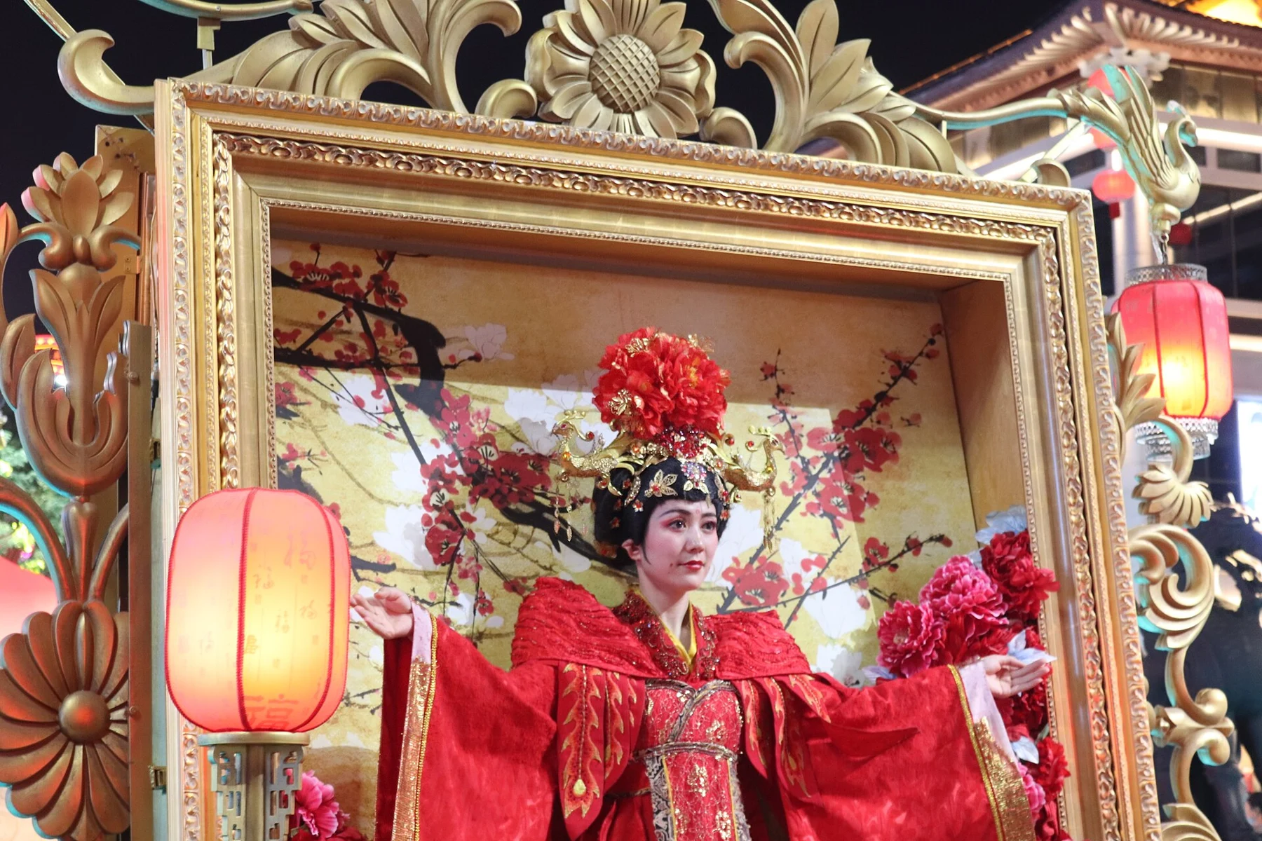 A red-costumed performer within an ornate rectangular display framed by gold decoration and lanterns.
