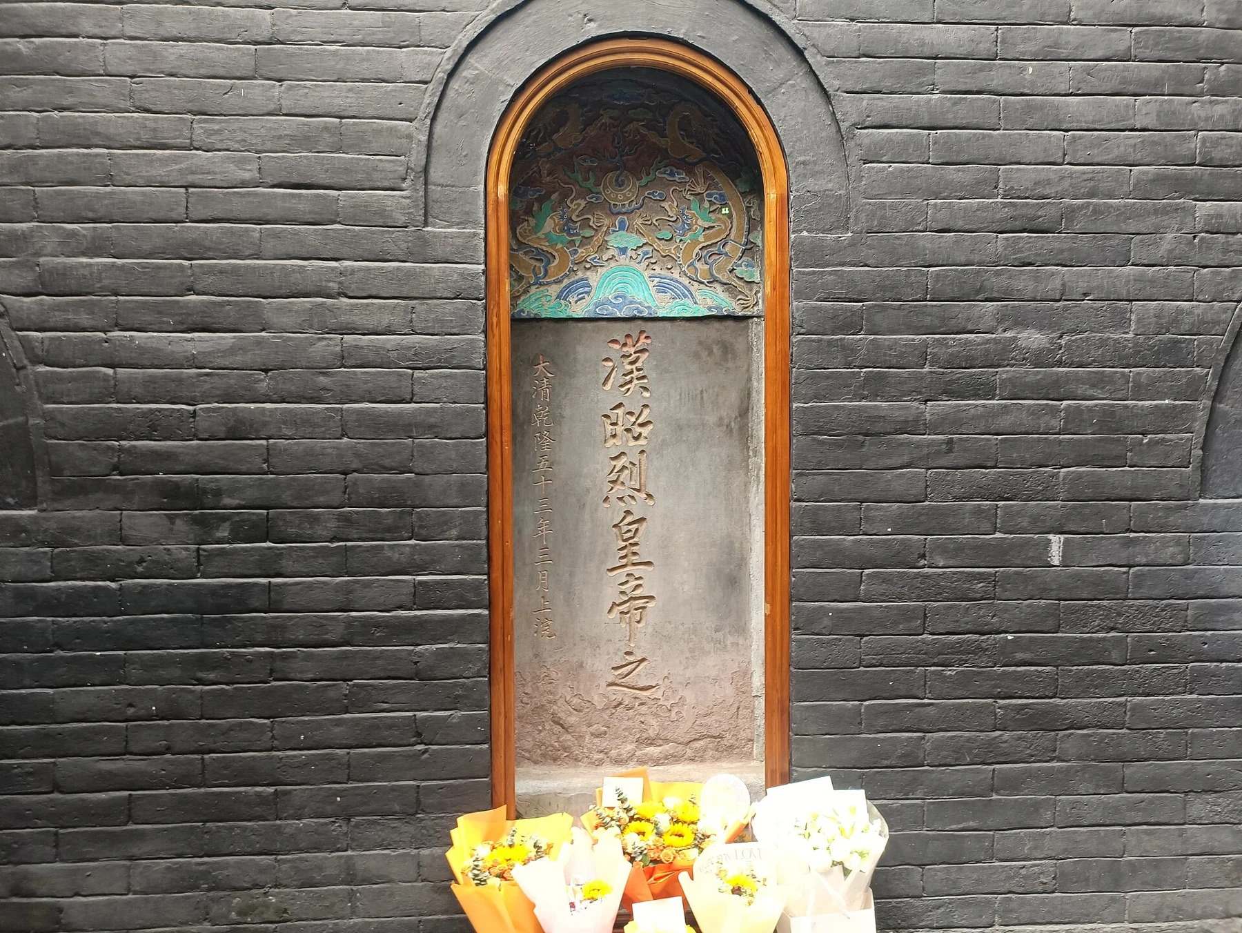 Inscribed memorial stone in an arched brick niche with flowers below