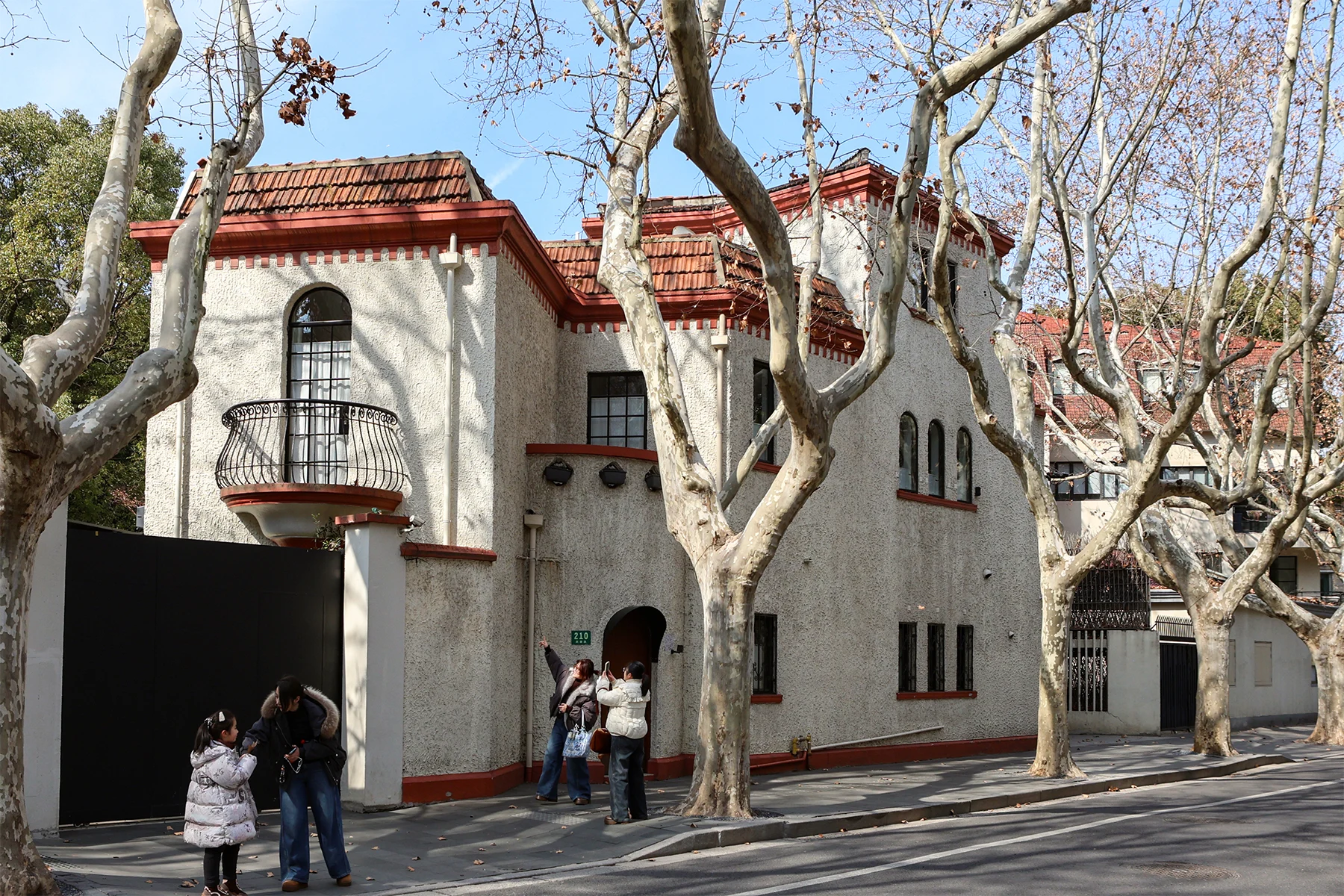 A pale low-rise building with red-tiled roofs, small curved balconies and bare tree trunks outside.