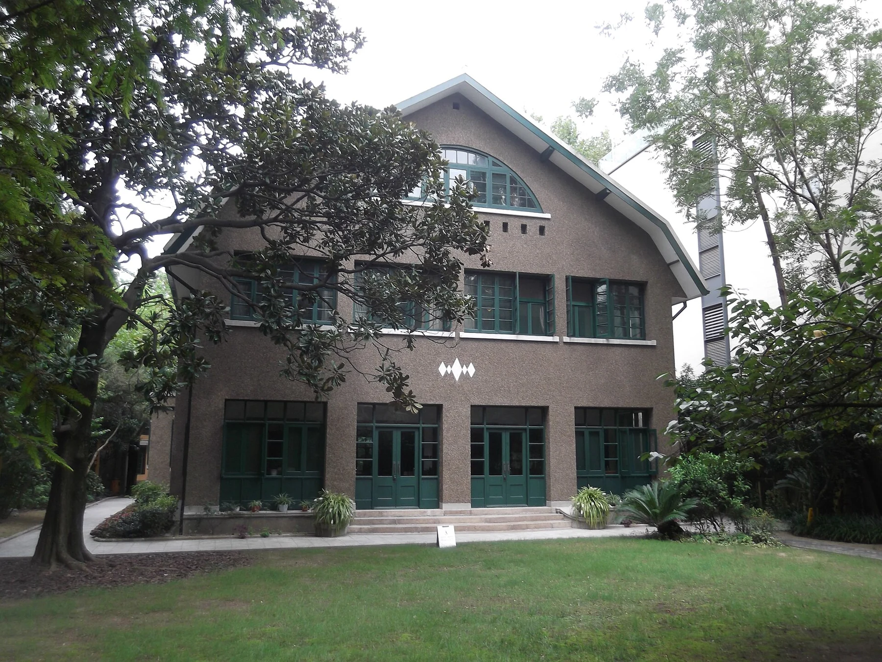 A pale house with green shutters and doors behind a lawn and garden trees.