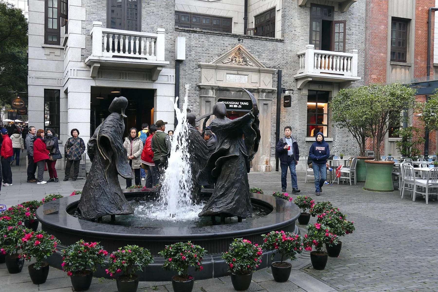 Three dark fountain figures with water, flowers and grey façades behind.