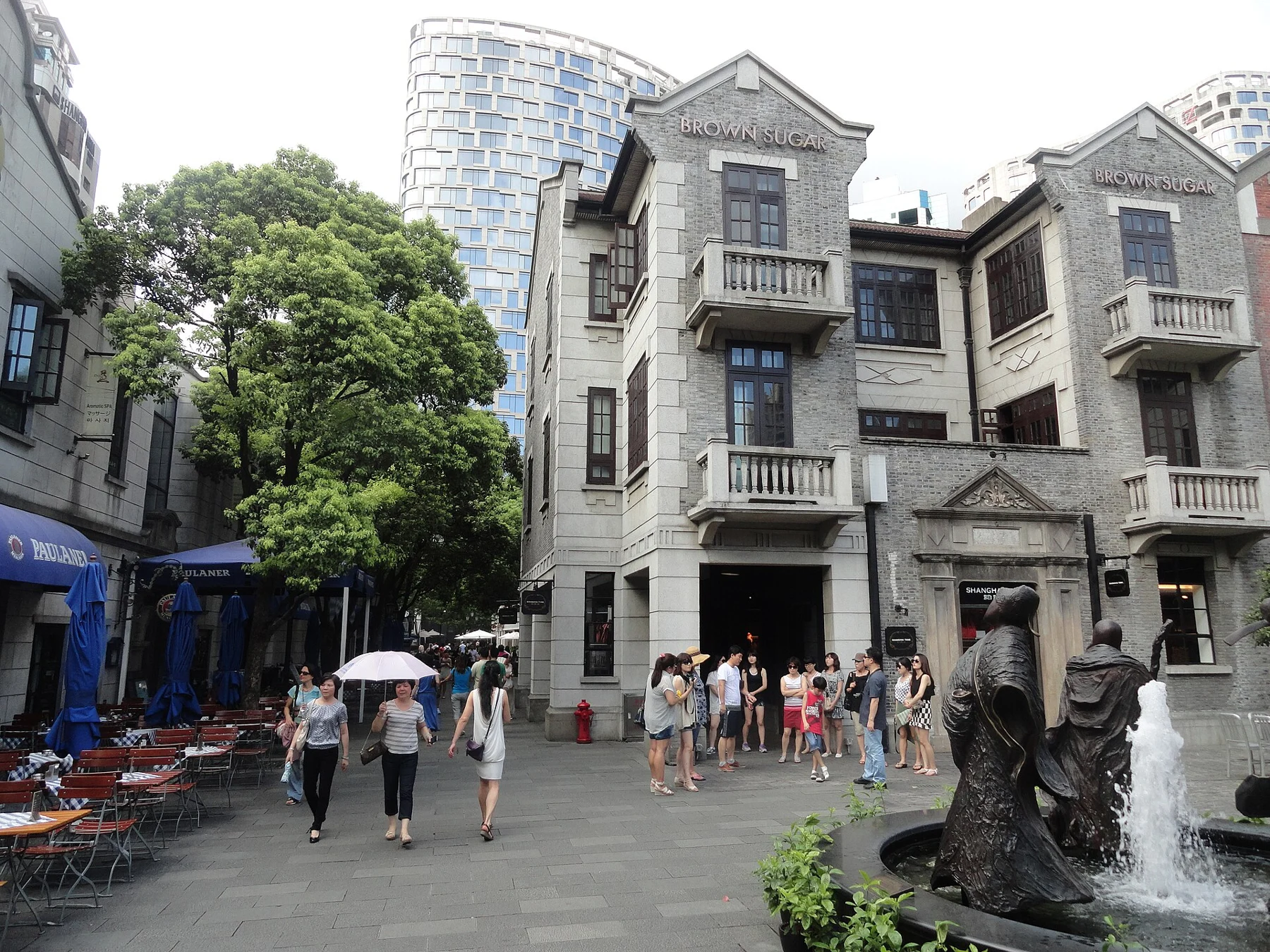 Grey low-rise façades with projecting balconies beside fountain sculptures and a plaza.