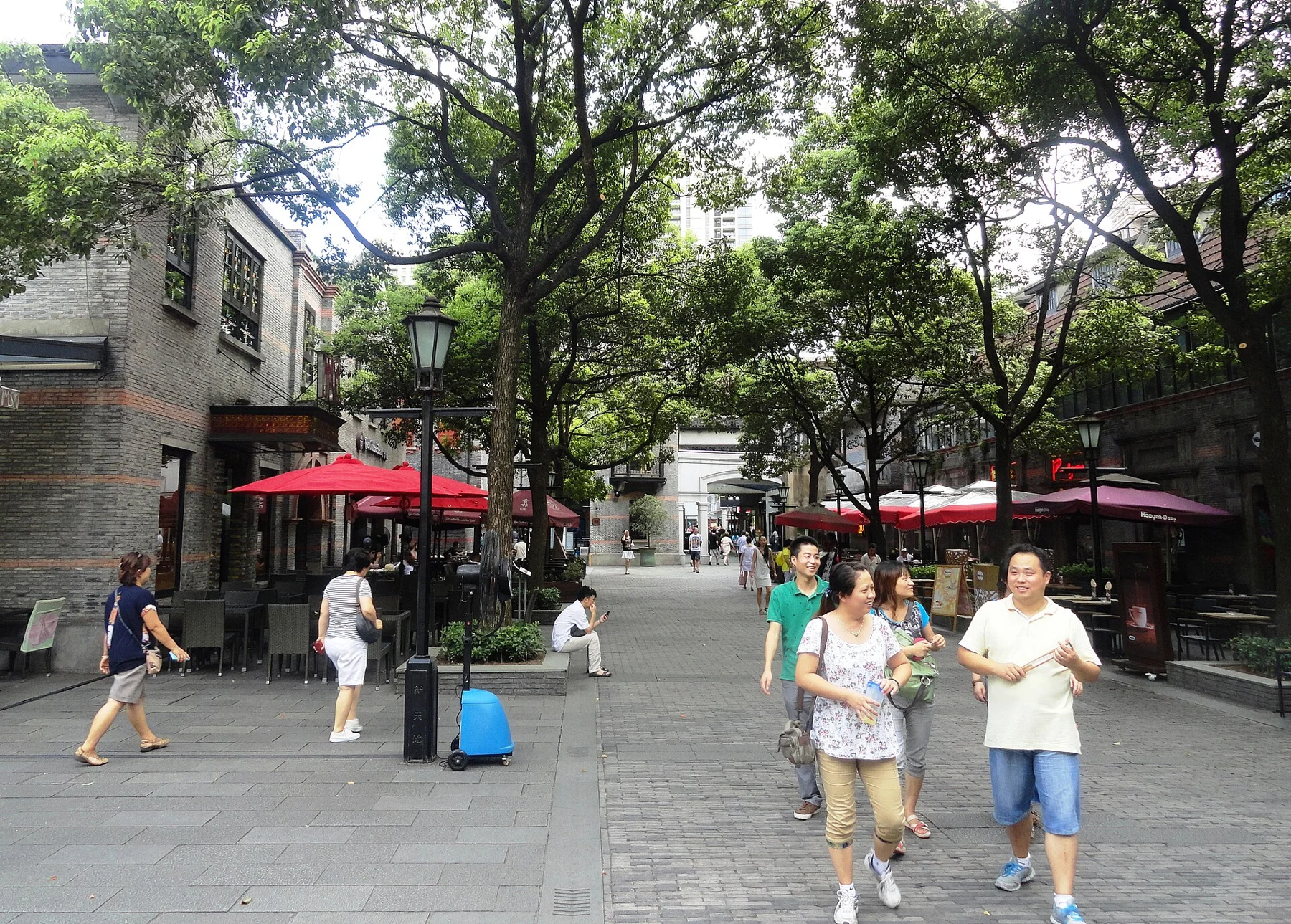 A pedestrian plaza surrounded by low shikumen-style façades, trees and café umbrellas.