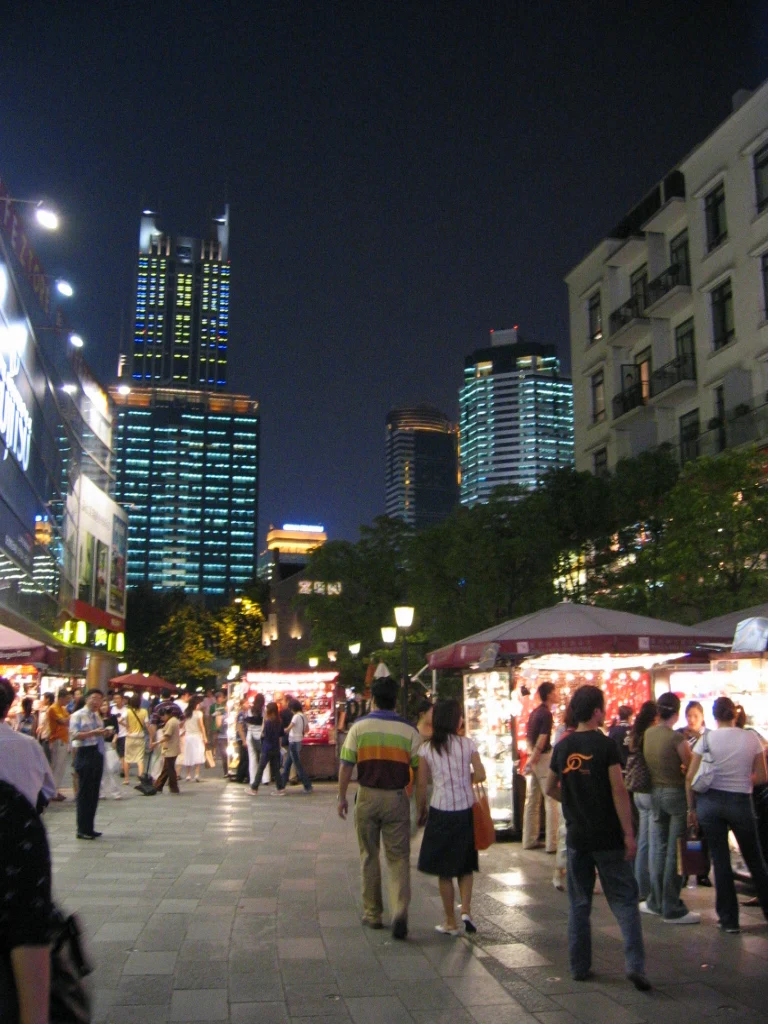 A lit pedestrian street at night between low façades, with visitors and taller illuminated buildings beyond.