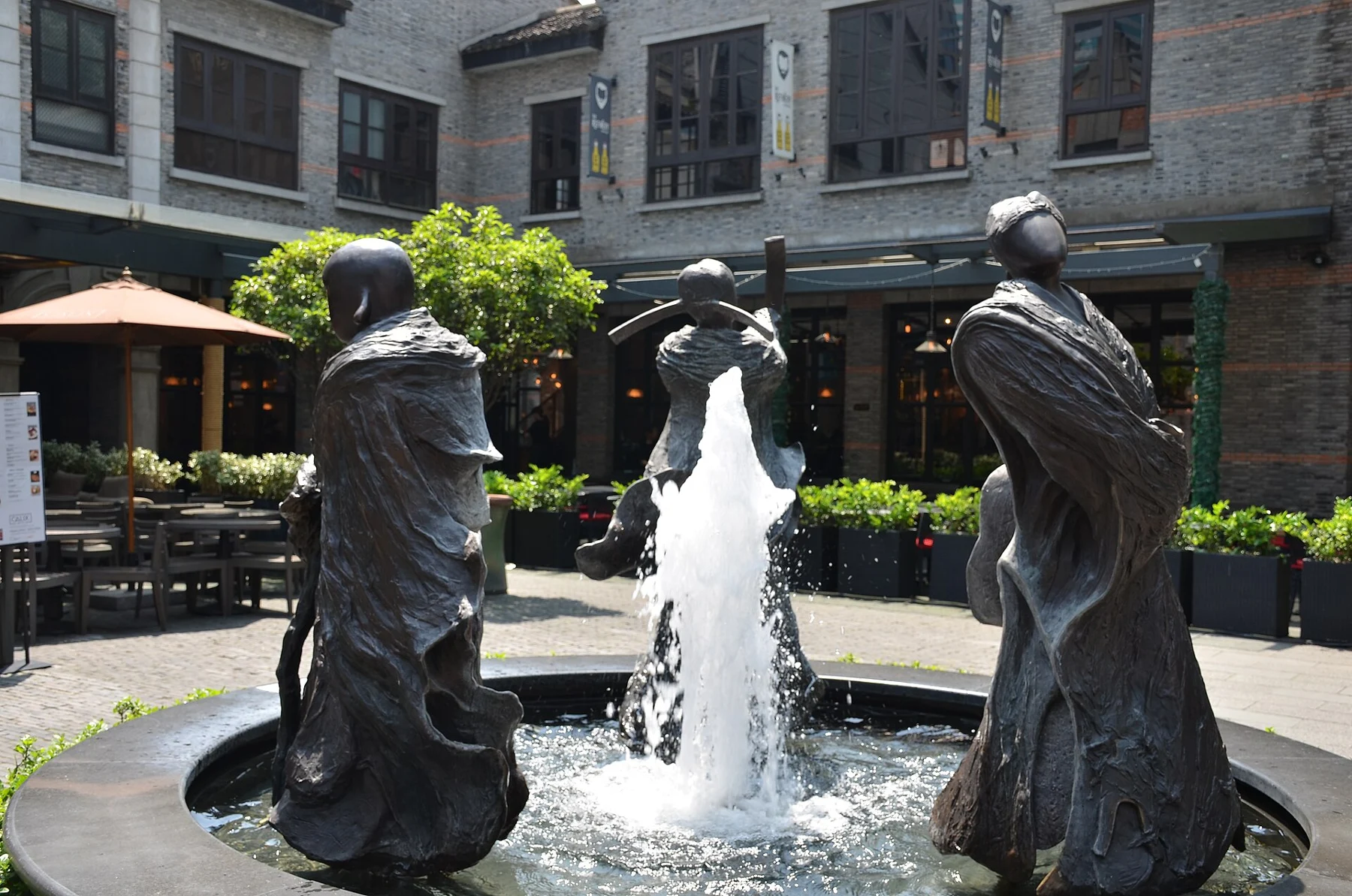 Three dark standing figures around a jet of water, with café tables behind.