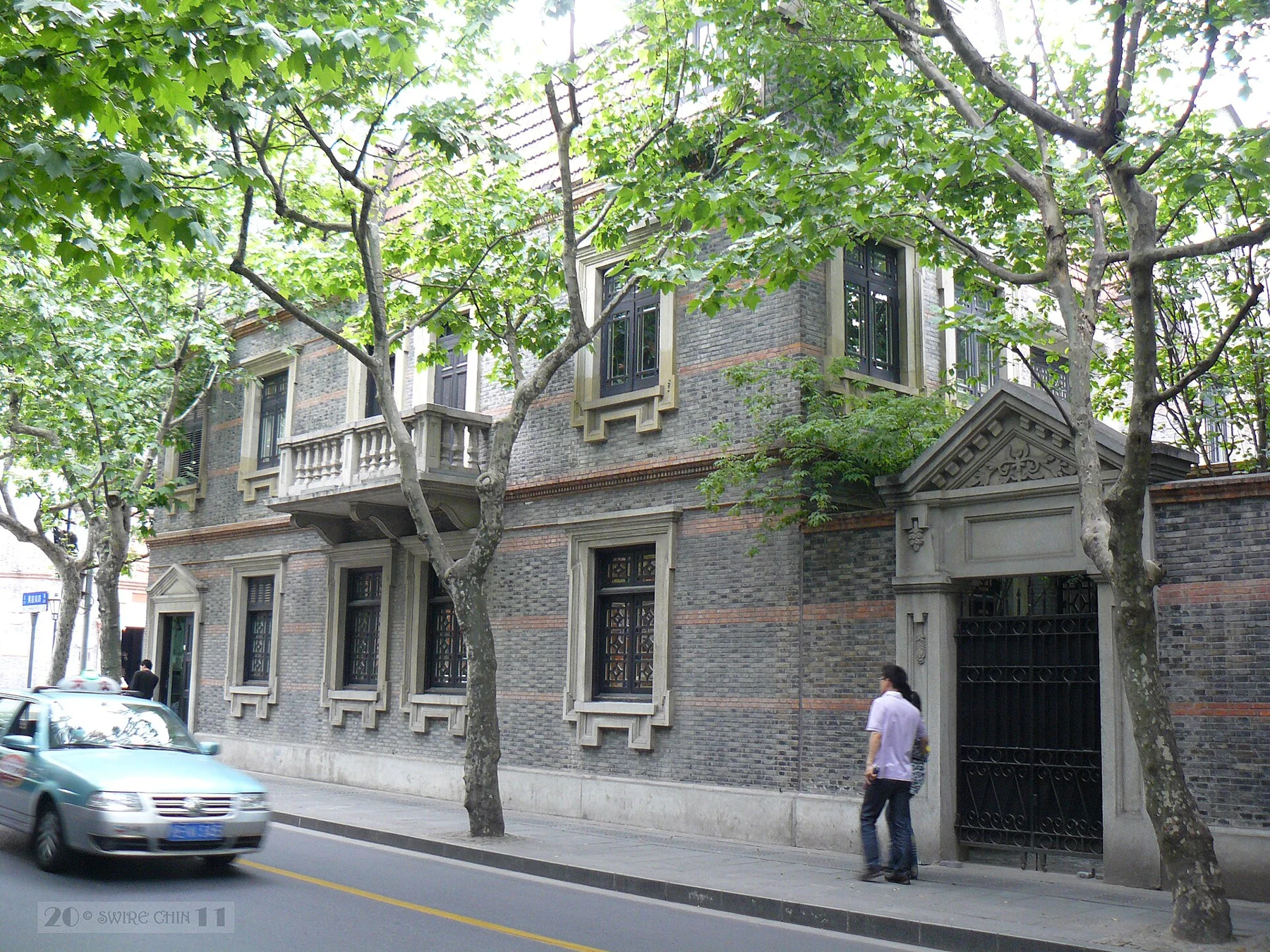 Stone-framed doorways set into grey brick façades, with trees and a car on the street.