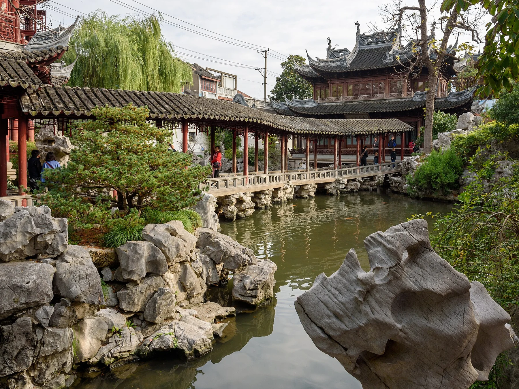 A red-columned covered corridor bending along the pond with rocks and a tower beyond.