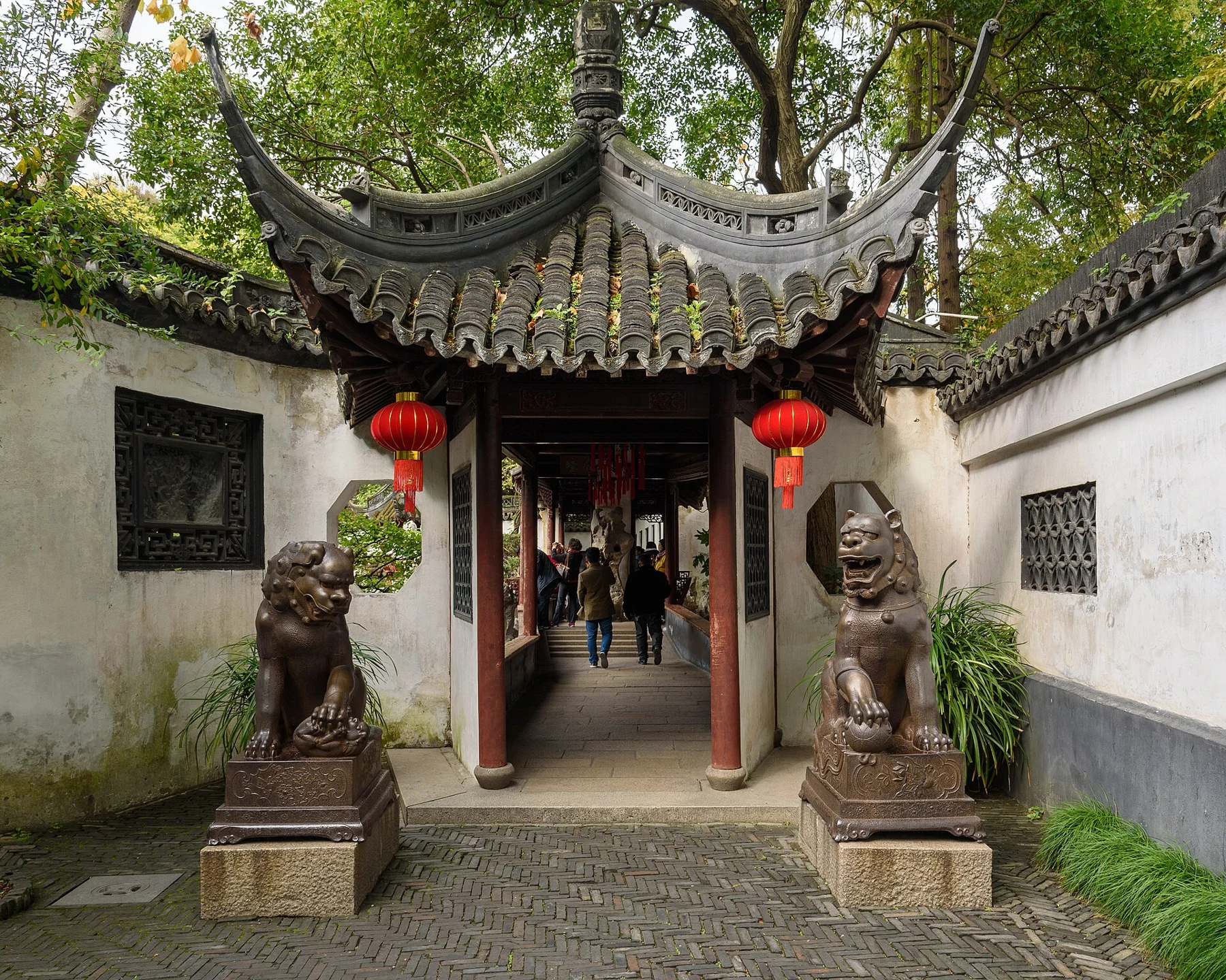 Two dark metal lion figures on plinths flanking the approach to a small roofed corridor.