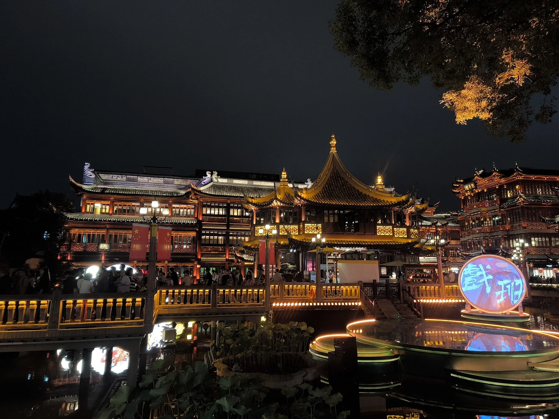 Visitors and illuminated traditional-style buildings around the bridge and pond at night.