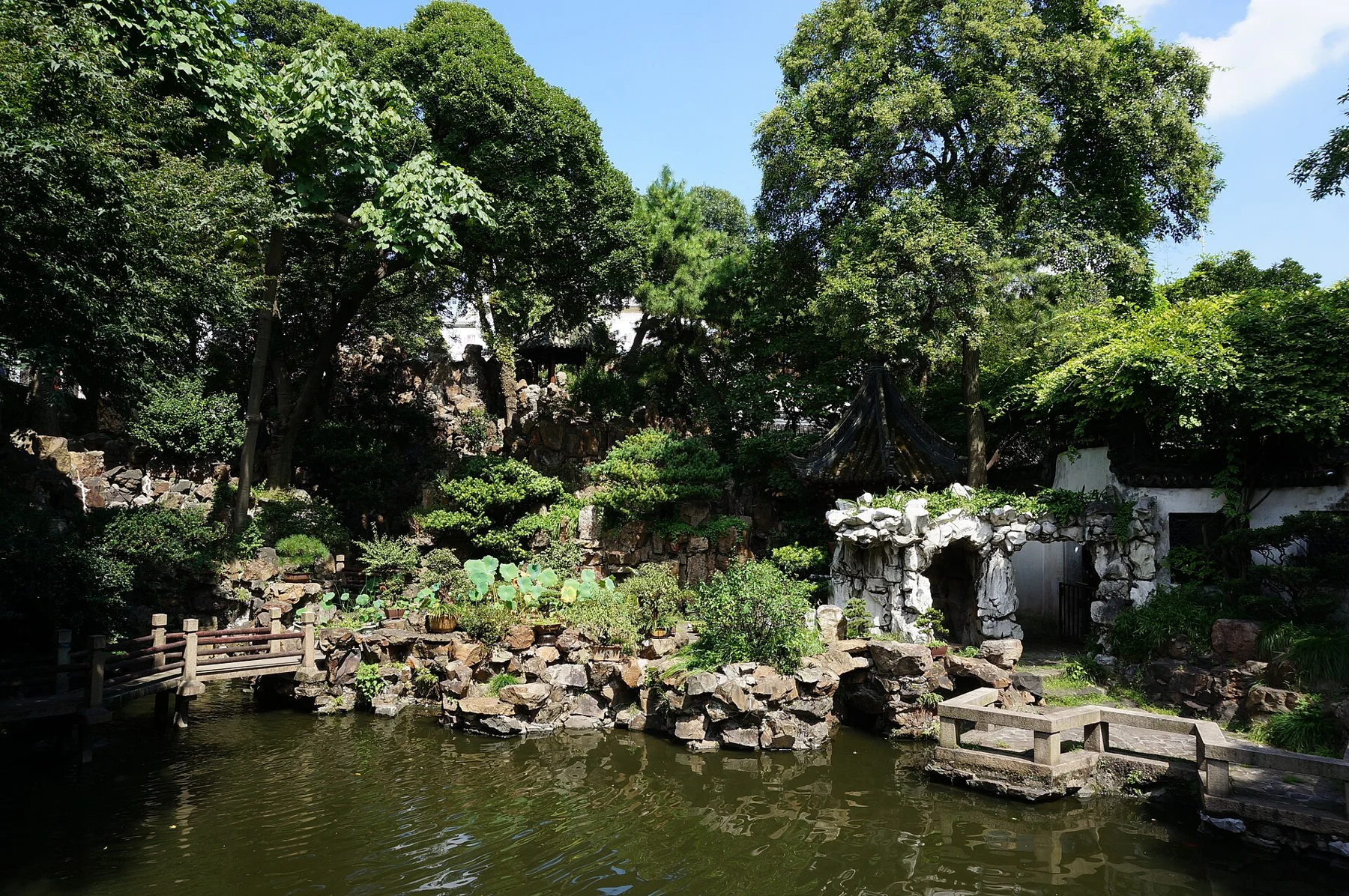 A pond and stone bridge below wooded rockwork, with a small pavilion and pale rock gateway.