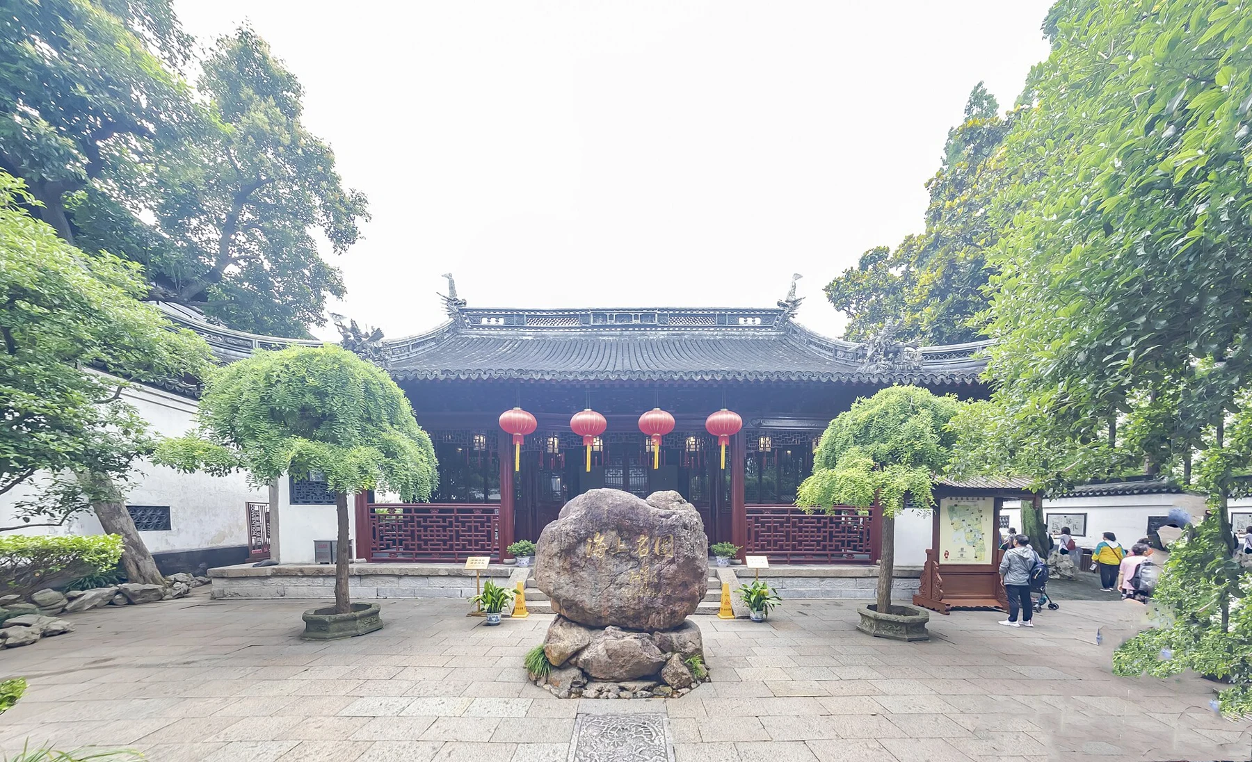 A red-and-dark timber hall behind a paved courtyard, an ornamental stone and trees.