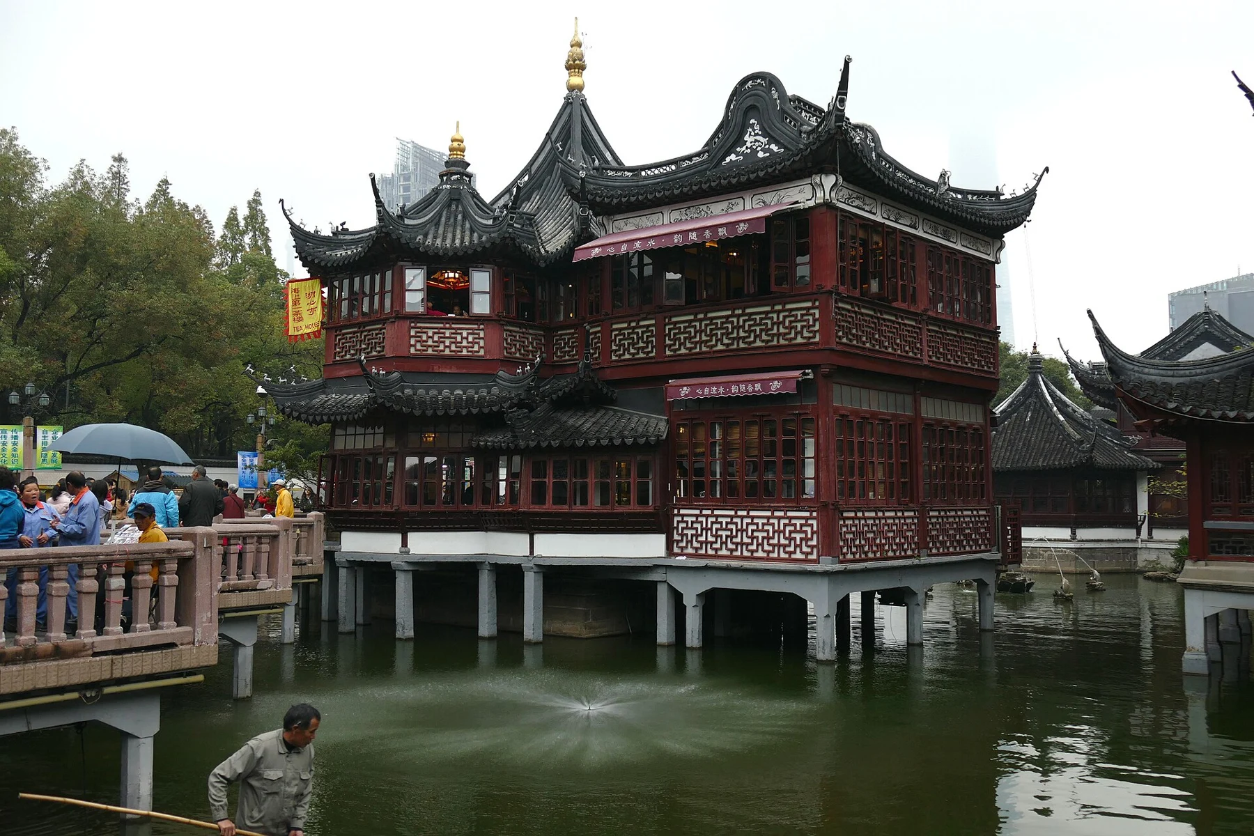 A red timber teahouse standing over a pond, with a worker in a small boat in the foreground.