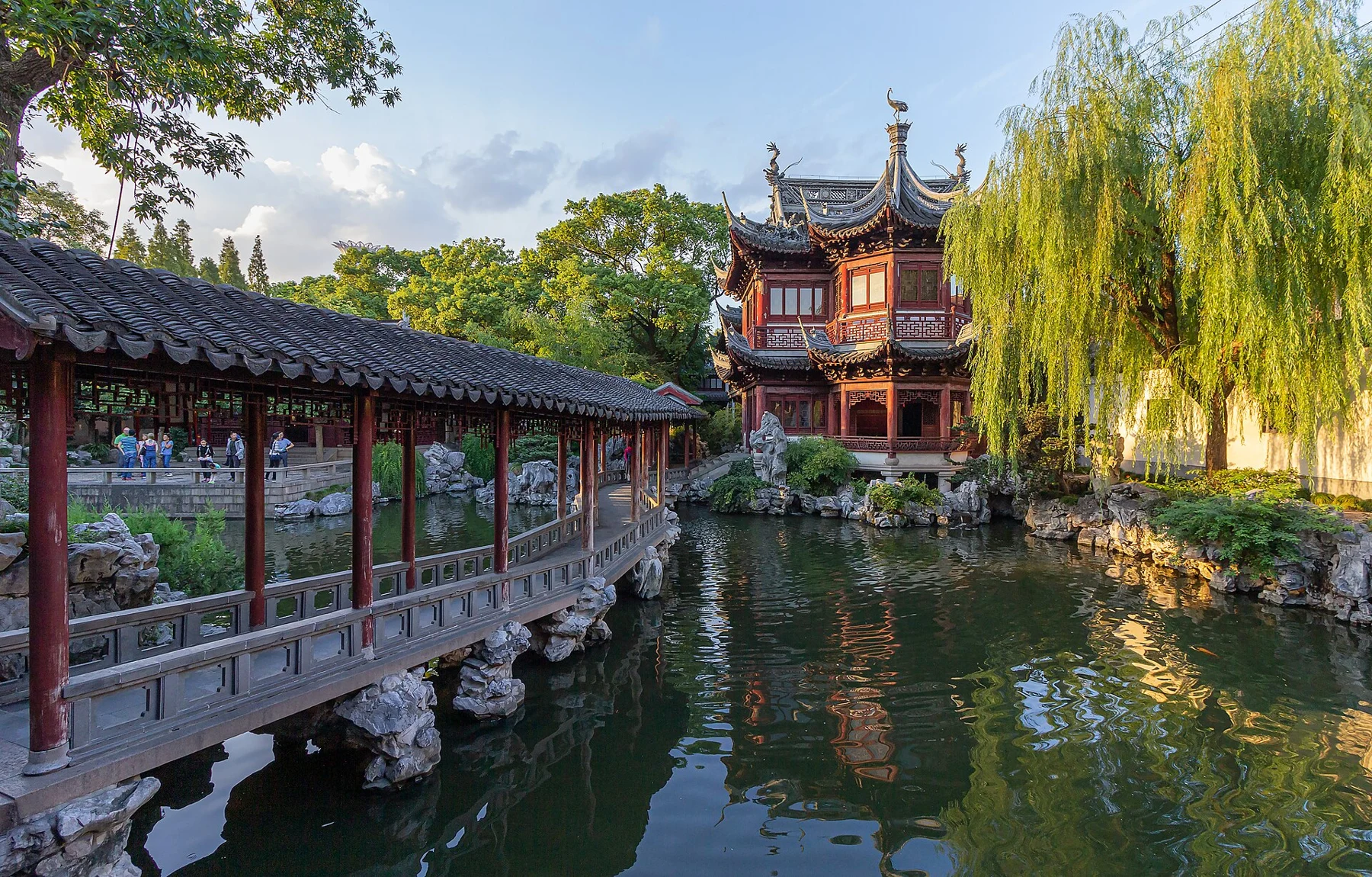 A red timber tower, covered corridor, willow and pond reflecting the garden buildings.