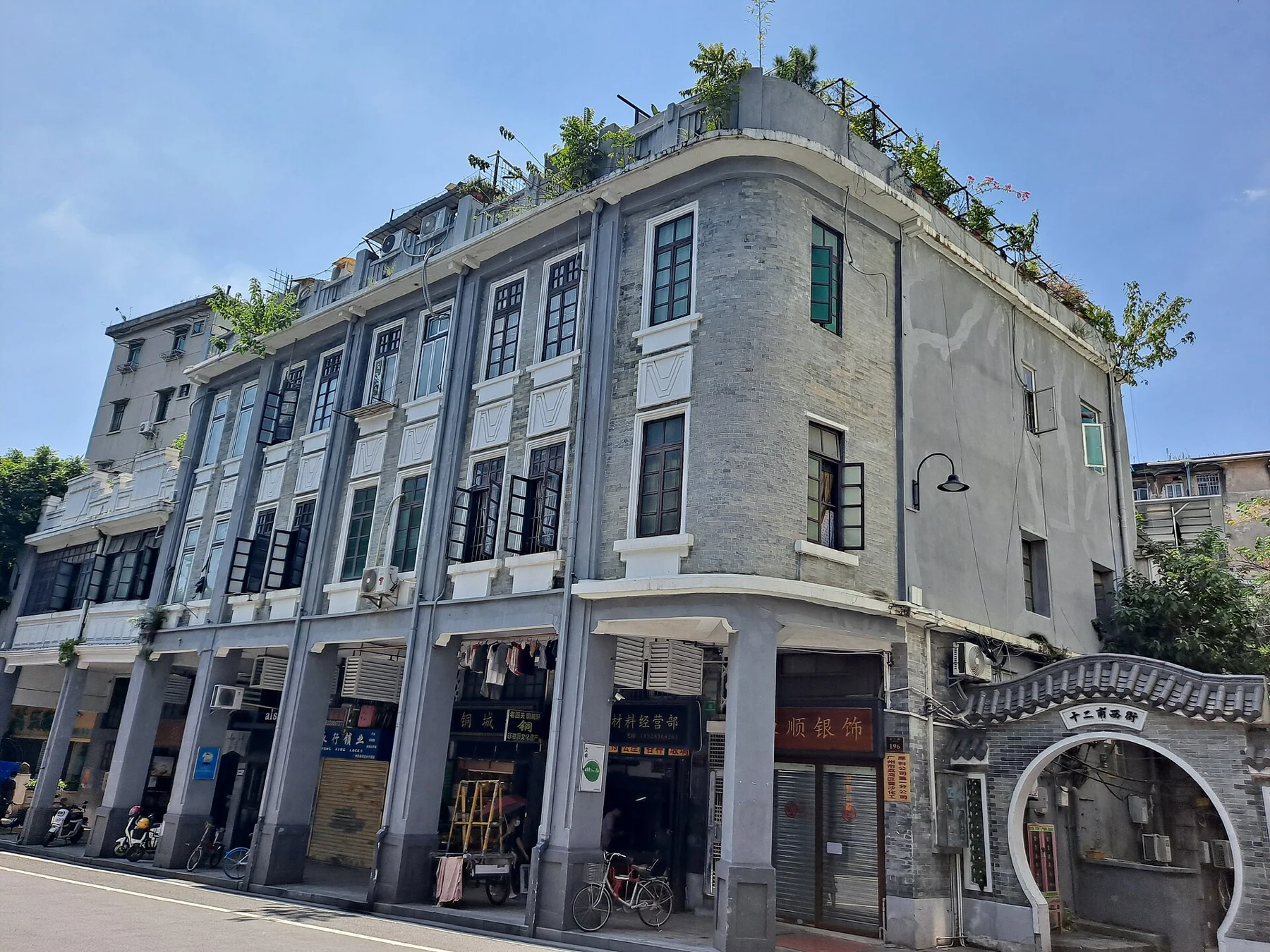 Grey corner building with tall windows, an arcade and an adjoining arched entrance.
