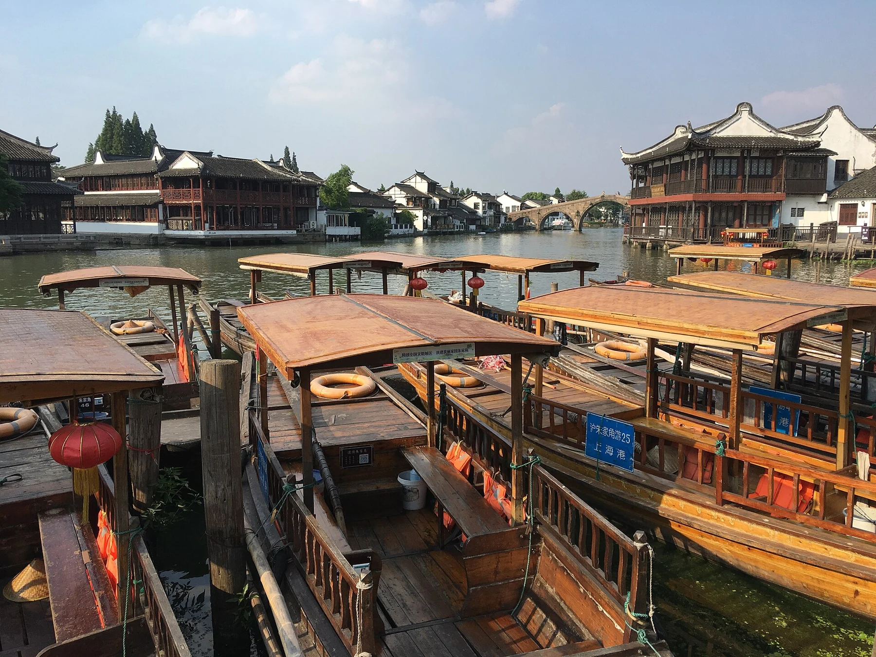 Roofed wooden boats at a dock, with riverside buildings and distant stone arches.