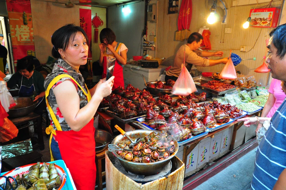 People beside trays of prepared food, leaf parcels and hanging shop lights.