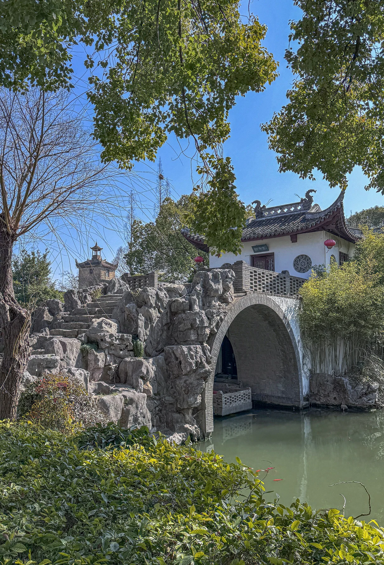 A small arched bridge over water beside rockwork, trees and a white-walled building.