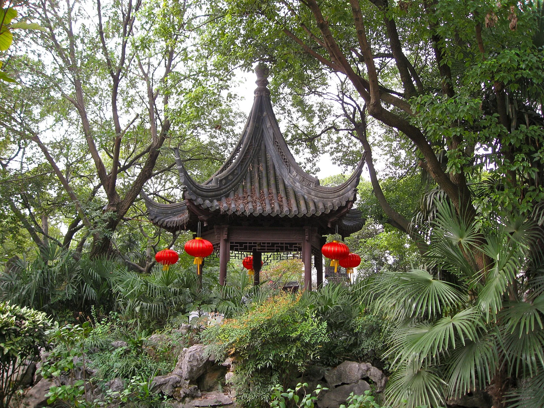 A pointed-roof pavilion with red lanterns surrounded by trees and rocks.