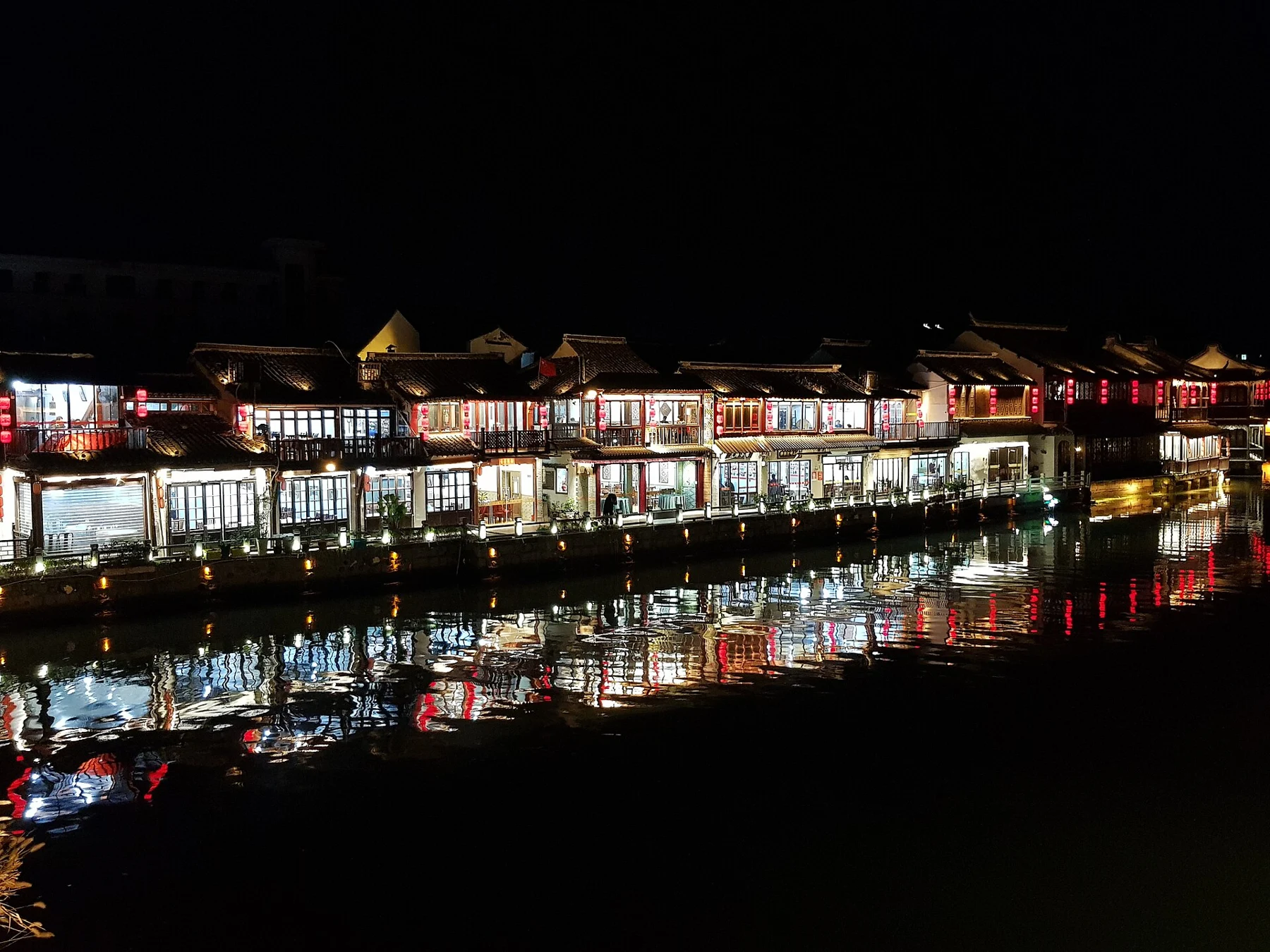 Lit shopfronts and tiled roofs reflected in dark water.