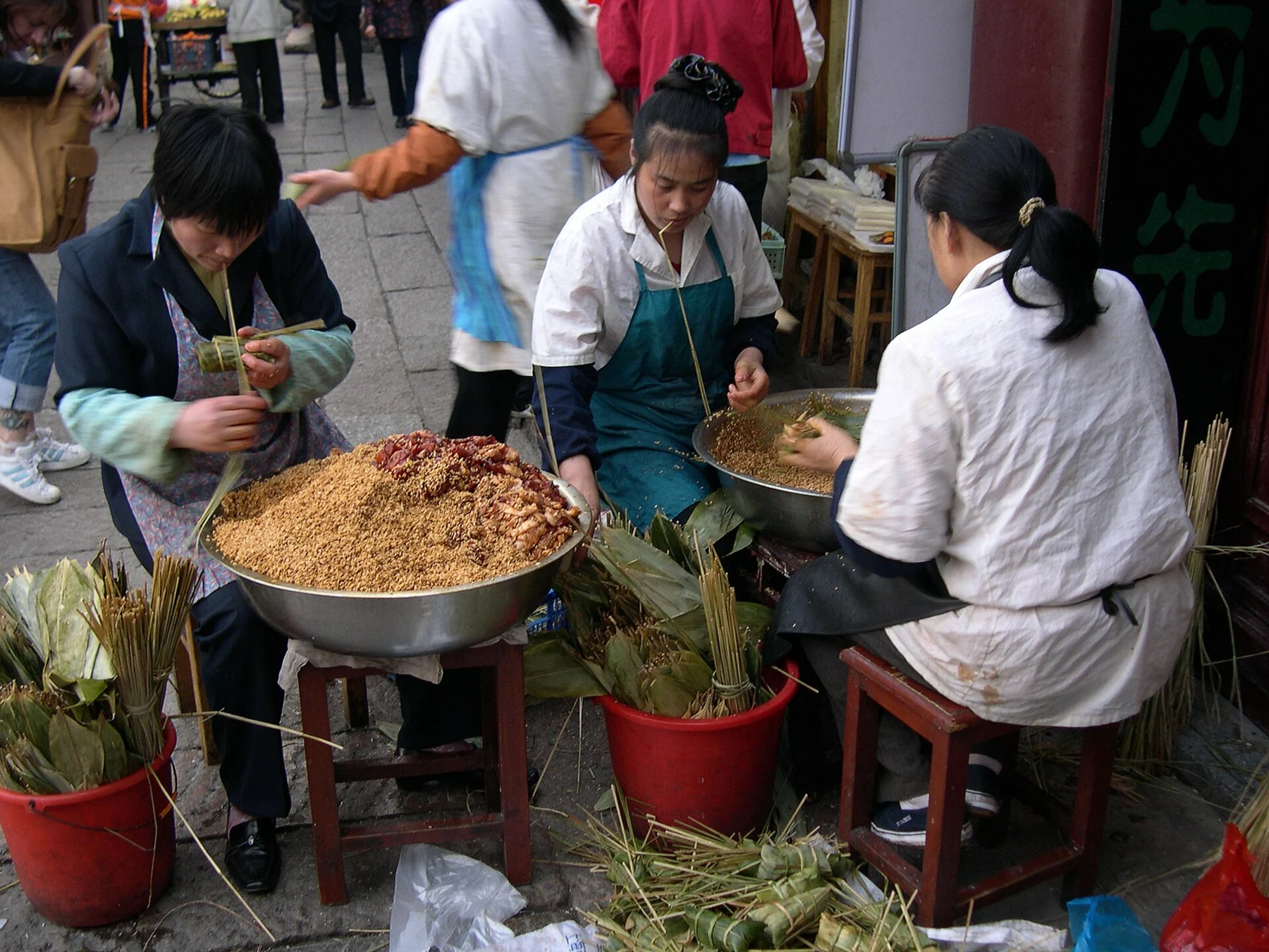 Women seated at a small work area using leaves and rice to prepare wrapped parcels.