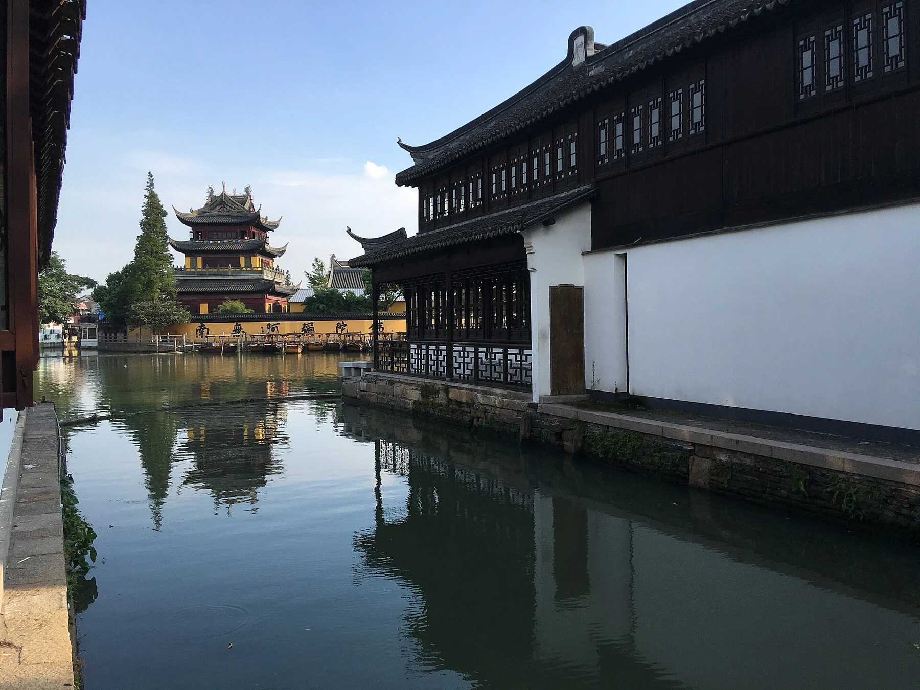 A canal reflecting a long white wall and dark timber upper storey, with a pavilion beyond.