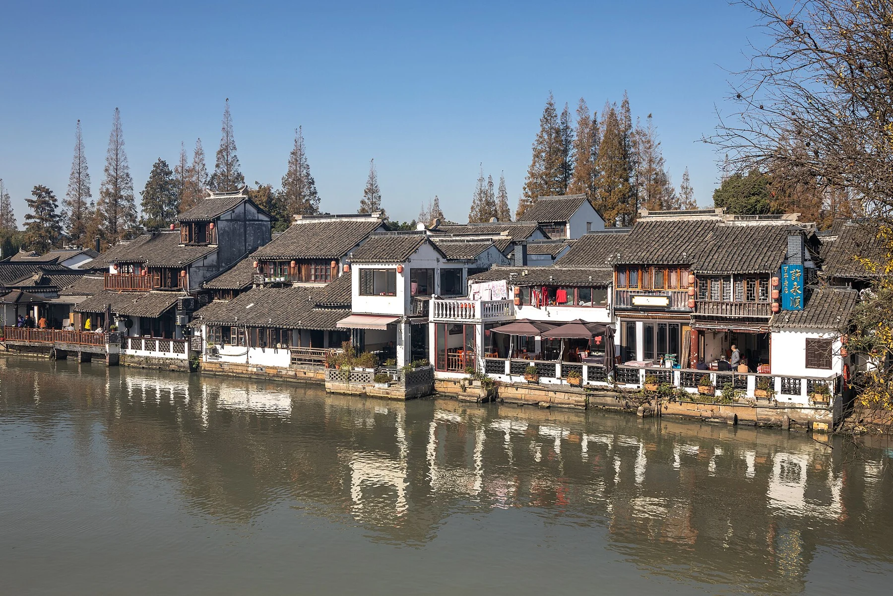 Rows of tiled-roof buildings reflected in calm water beneath trees.